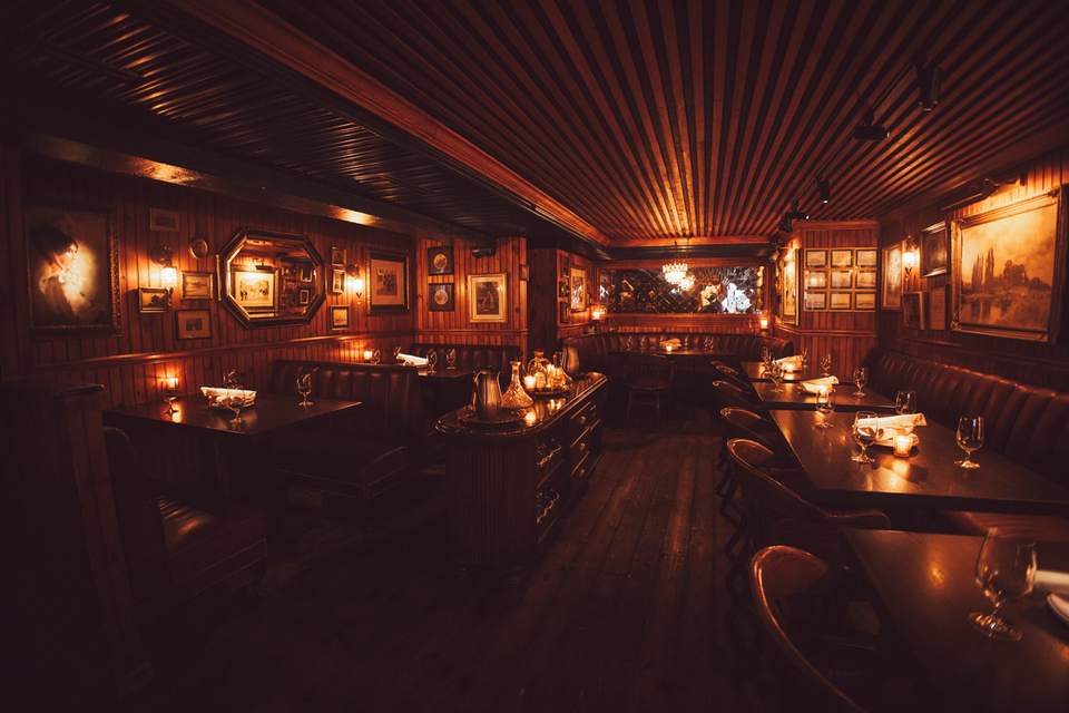 Diners at a dimly lit table in a restaurant, with a large seafood tower, wine glasses, and a cocktail, against dark wood-paneled walls with framed
