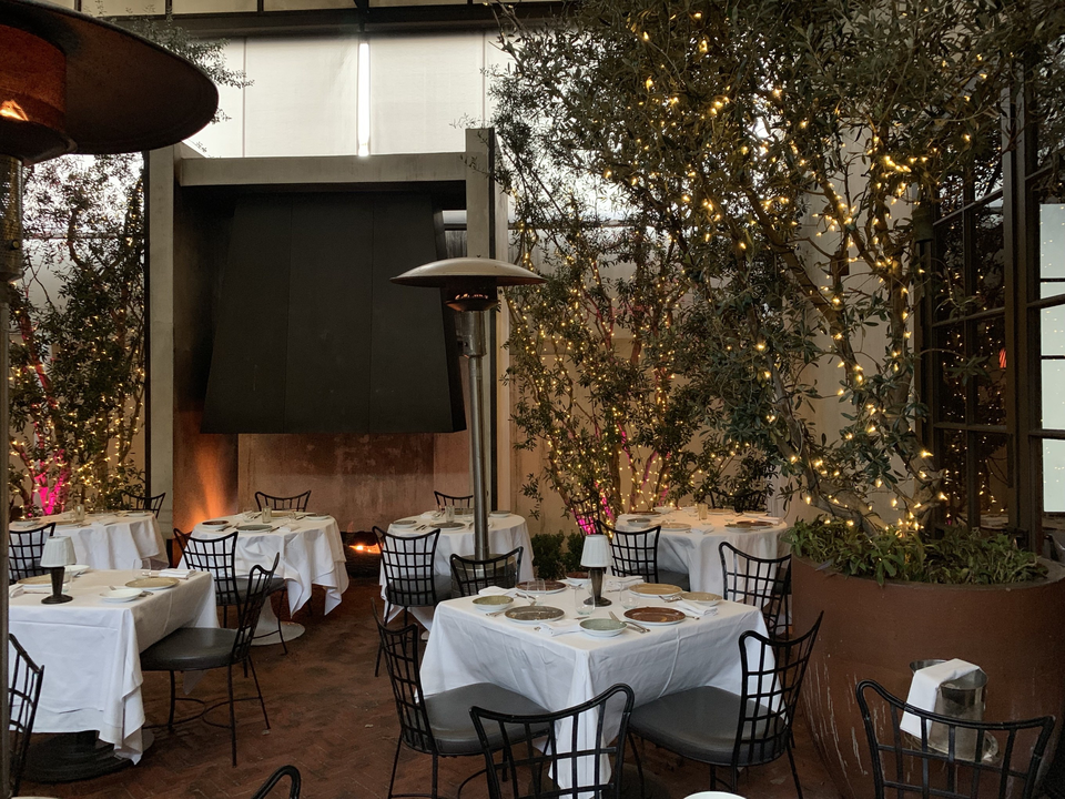 A restaurant dining room with light wood tables, black chairs, pendant lighting, a wine wall, and bar seating.