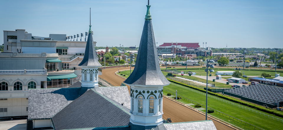 Aerial view of Churchill Downs racetrack showing white buildings with teal-roofed twin spires, manicured grounds, and horse racing facilities.