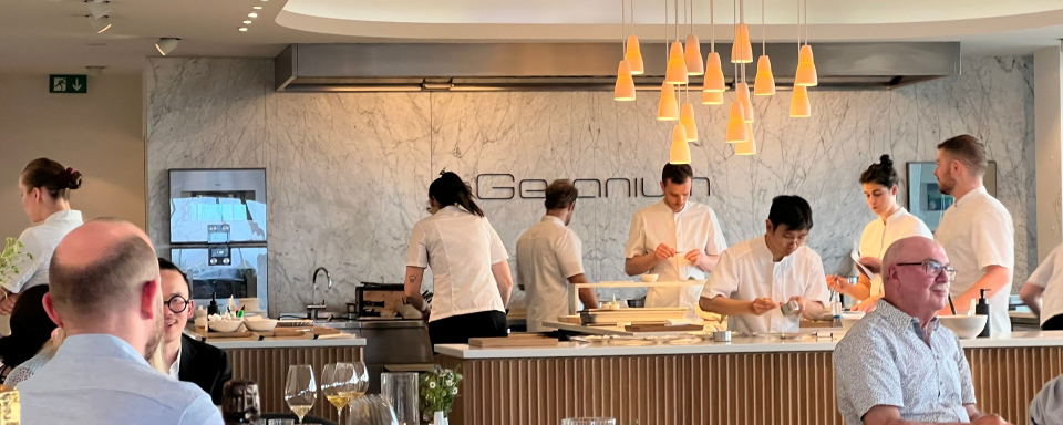 The interior of Geranium Restaurant shows chefs working at an open kitchen counter with a marble backsplash and a staff member setting a table.