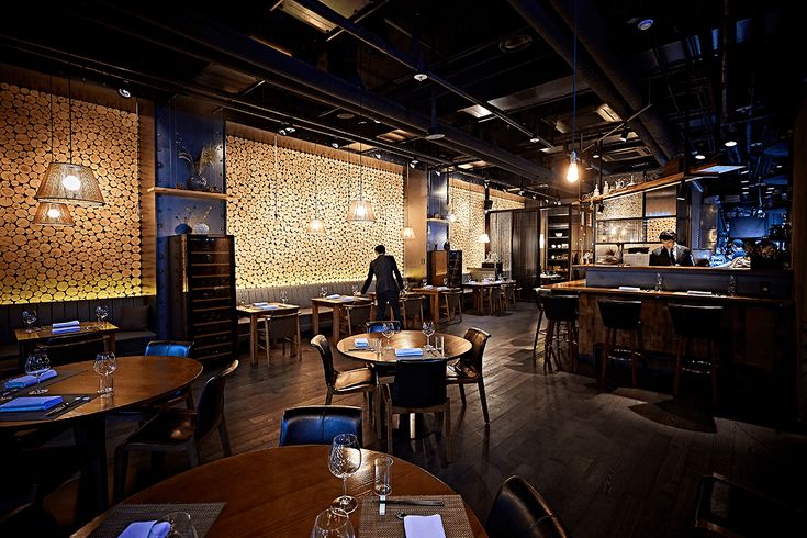 An upscale restaurant interior with wooden tables, black chairs, glowing stone wall panels, and a bar area visible in the background.