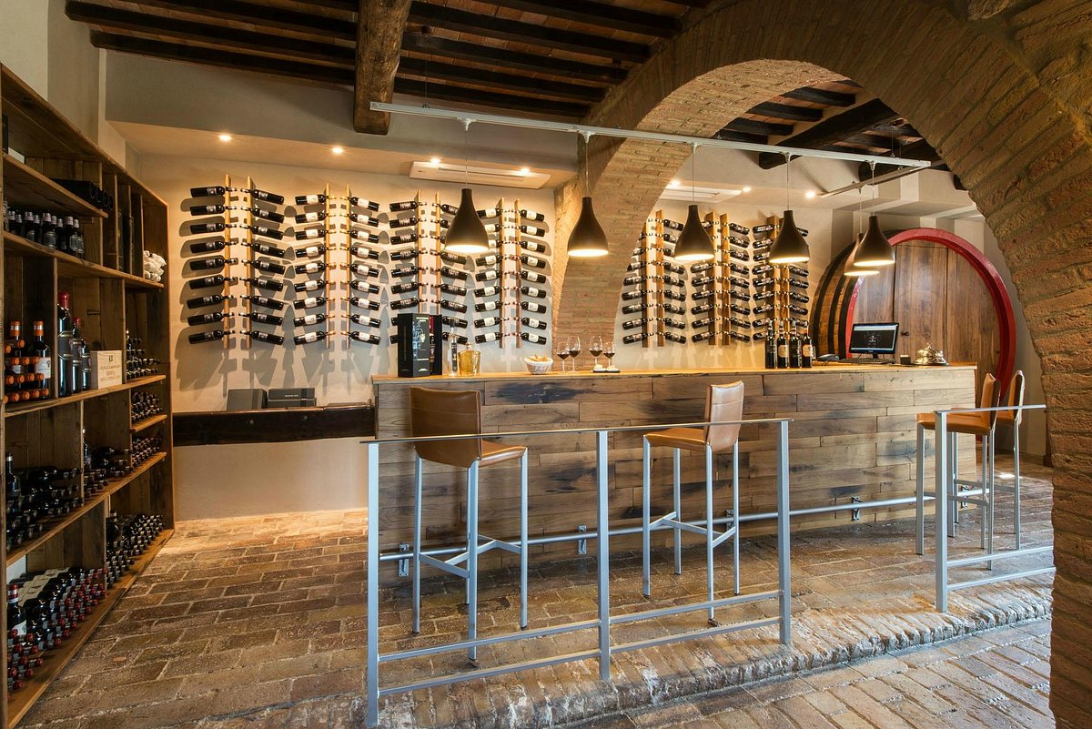 Interior of a wine tasting room with illuminated wine bottle racks on walls, a wooden bar, metal bar stools, exposed brick, and wooden ceiling beams.