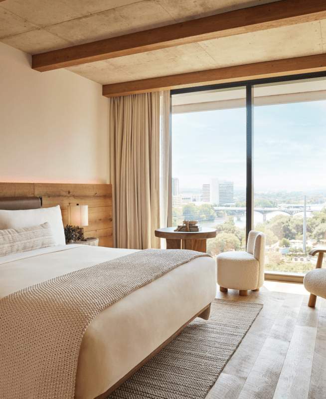 A modern hotel guest room with floor-to-ceiling windows overlooking a city skyline, featuring natural wood accents, oat-colored bedding and textiles,.