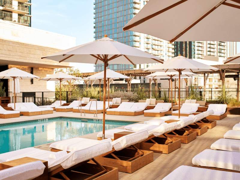 A luxurious rooftop pool area with wooden lounge chairs, white cushions, and tan umbrellas overlooking a swimming pool and city buildings.