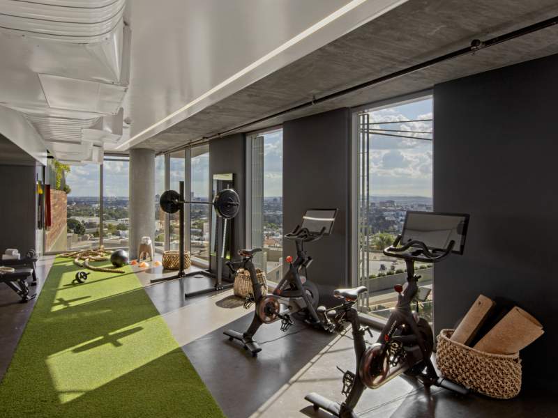 A high-rise gym with exercise bikes, large windows showing a cityscape view, green turf flooring, and dark walls with contemporary fitness equipment.