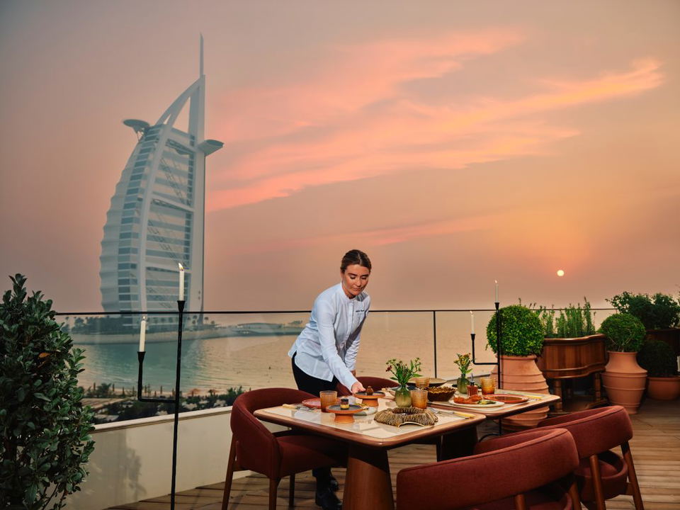 Outdoor terrace dining setup at sunset with Burj Al Arab visible in background, server setting table with Dubai skyline view