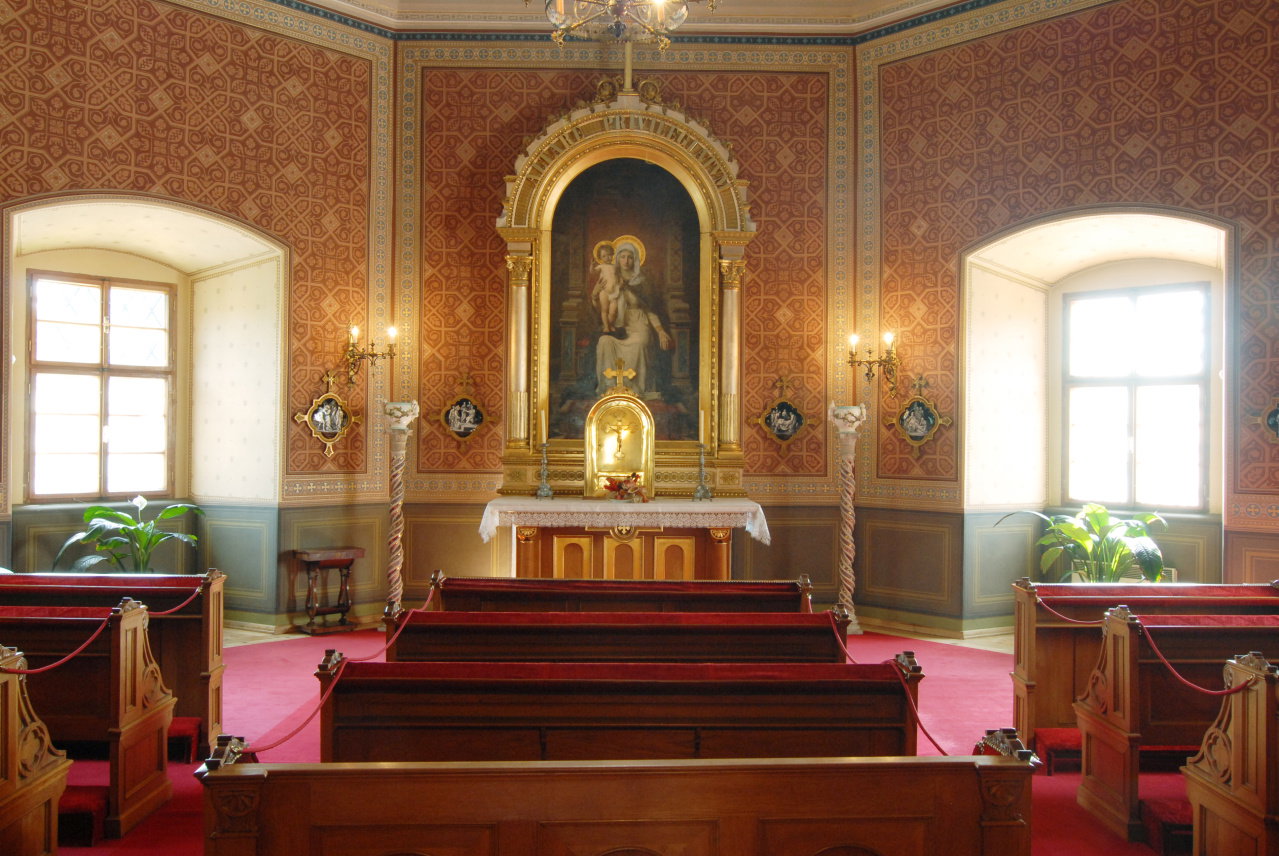 Interior of Bečov Castle chapel showing red carpeted pews, ornate patterned walls, and a gilded altar with a painting of the Virgin Mary and child.