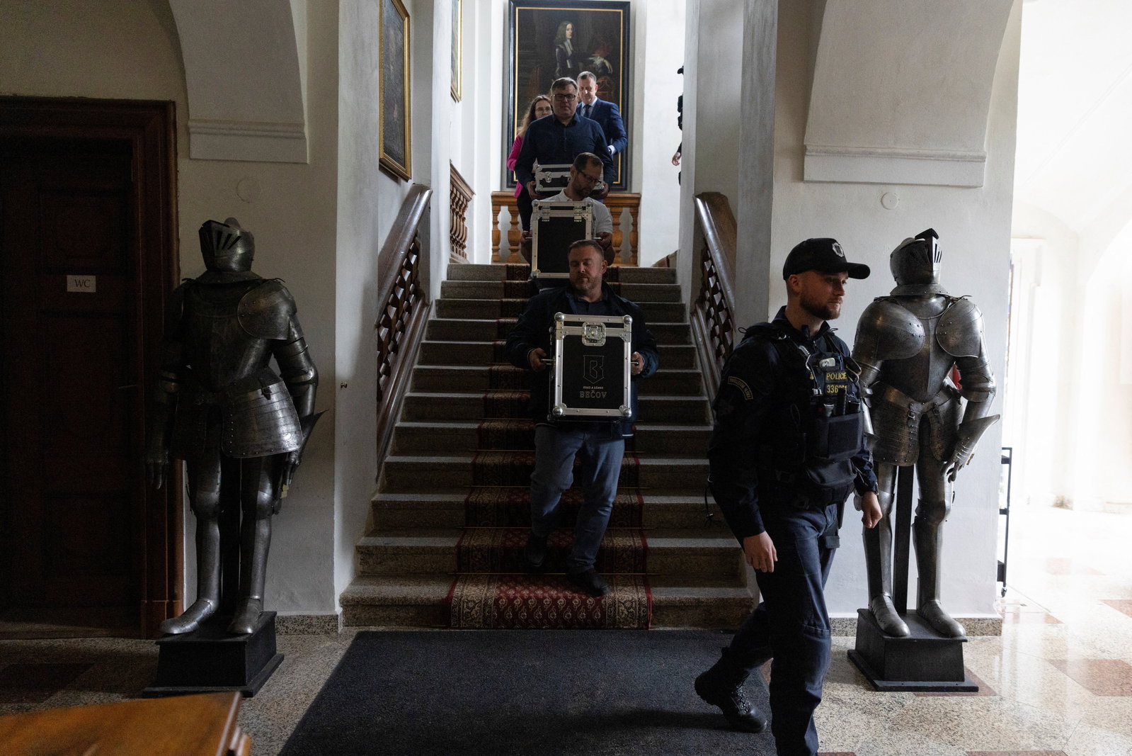A police officer and several men carry branded Becov Castle cases down a red-carpeted staircase flanked by two suits of armor, bearing restored 19th-century Chateau d'Yquem bottles.