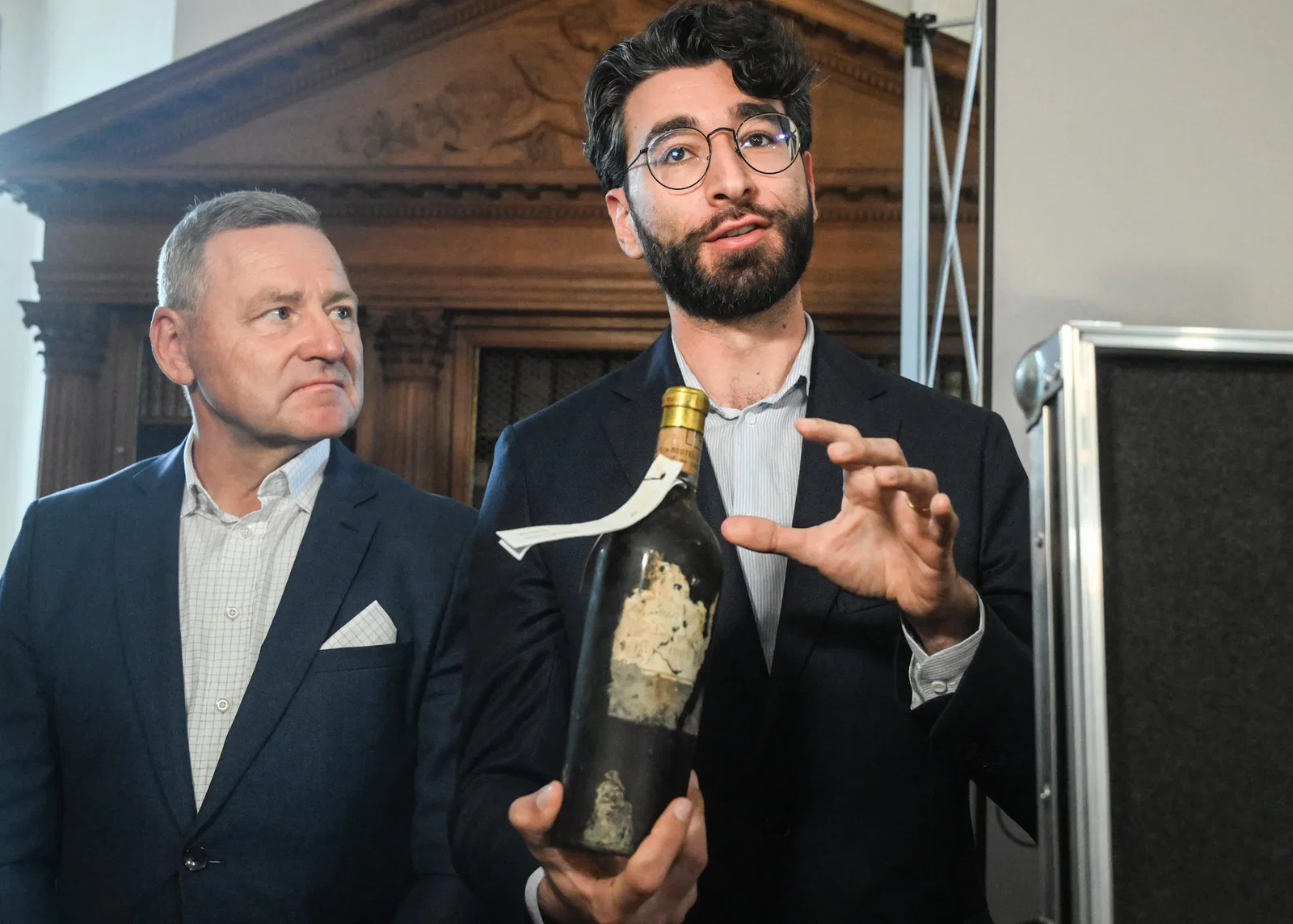 Two men in suits stand indoors, one holding an aged dark wine bottle with a deteriorated label and gold capsule, gesturing while speaking.