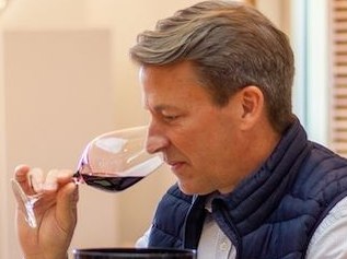 A man wearing a dark blue vest over a white shirt drinks red wine from a glass while appearing to assess its flavor.