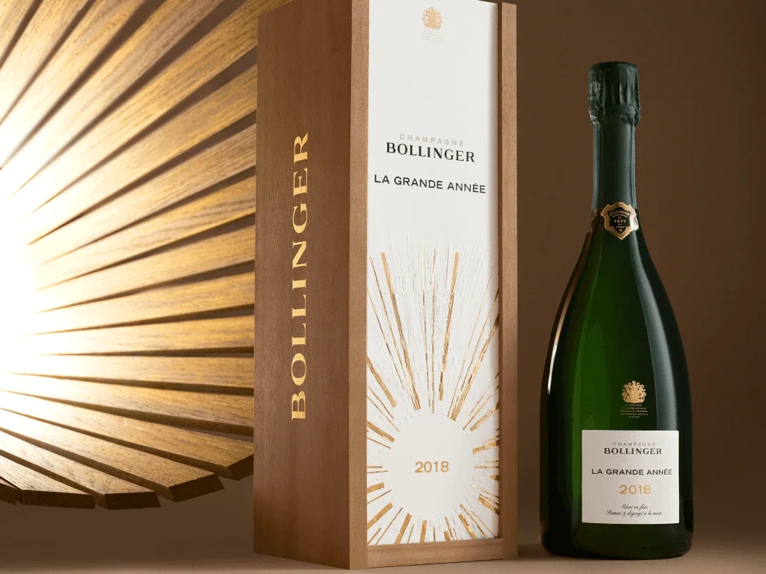 Bottle of Bollinger La Grande Année 2018 Champagne beside its wooden gift box with gold sunburst design, on a warm brown background.
