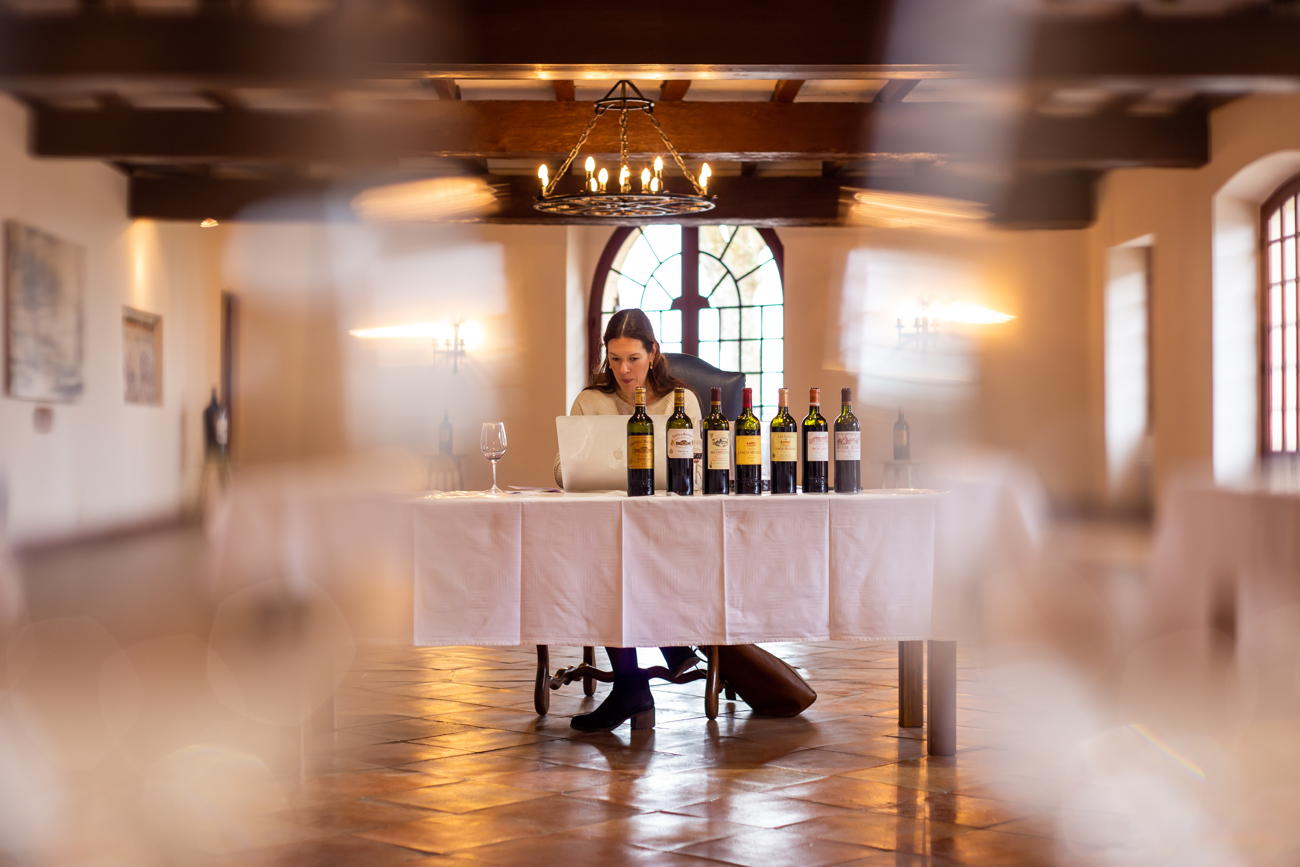 A woman sits at a white-draped table with seven wine bottles displayed in front of her, working on a laptop in a spacious room with warm lighting and architectural details.