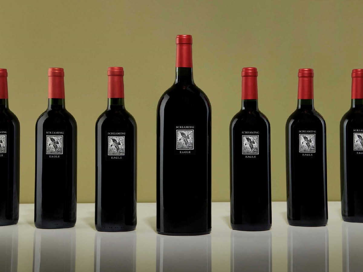 Seven dark glass wine bottles with red foil capsules and 'Screaming Eagle' labels, including one large-format magnum, on a white surface against an