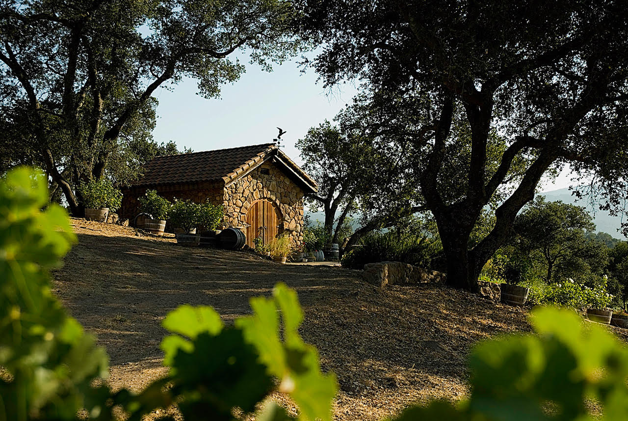 A stone winery building with a tiled roof and a weathervane, surrounded by trees and green foliage under a clear sky.