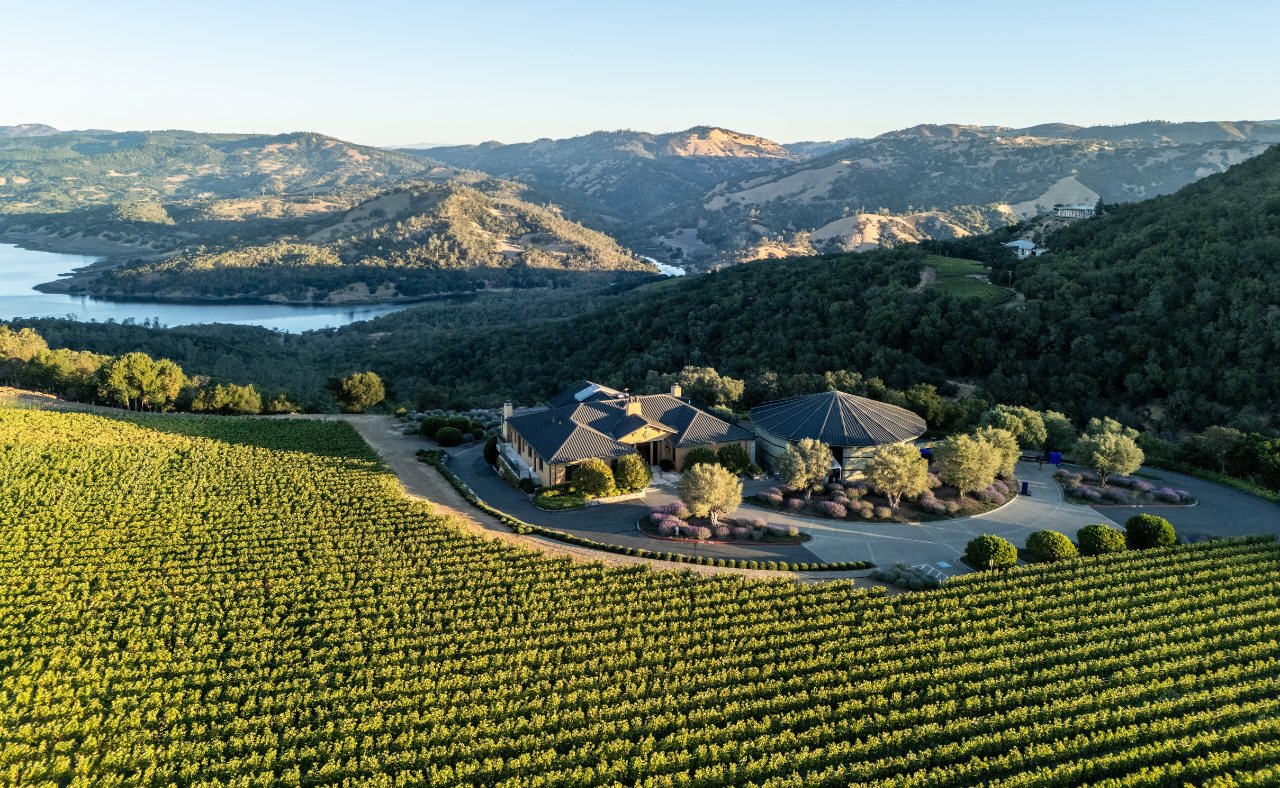 An aerial view of the Colgin Cellars estate, featuring a large, elegant building with a dark roof, surrounded by manicured lavender bushes and trees.