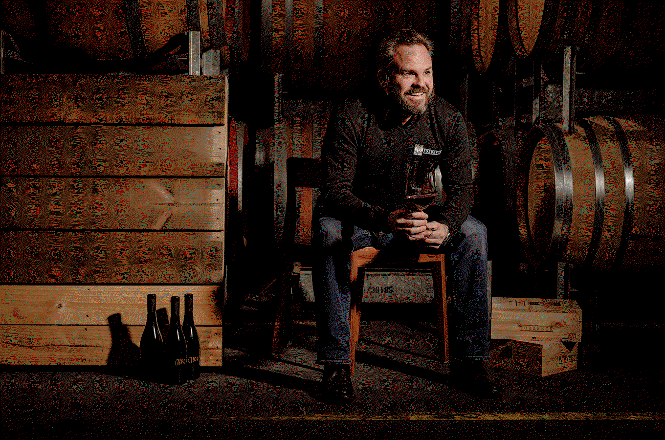 A winemaker sits in a barrel cellar holding a wine glass, with wine bottles visible in the foreground