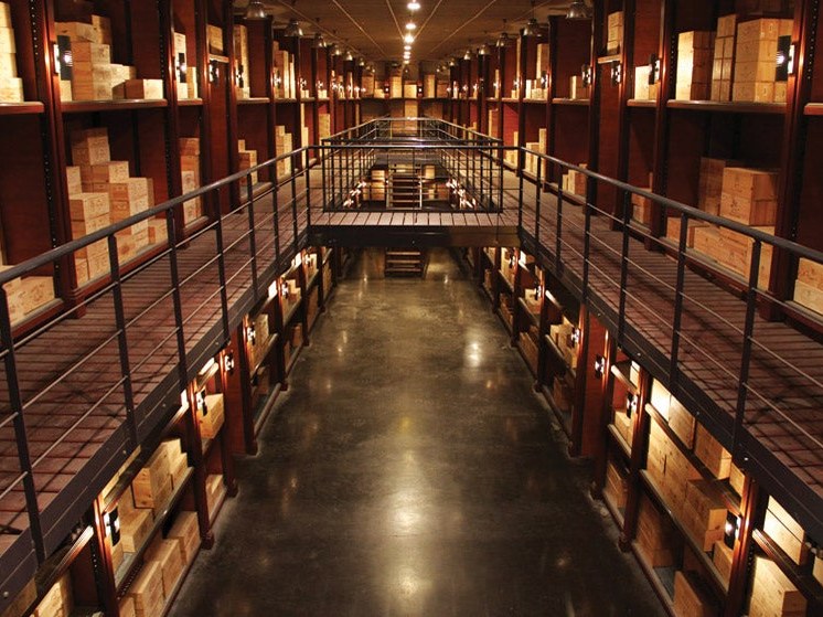 A multi-level wine storage warehouse with tall shelving units stacked with wooden wine cases, warm wall lighting, and a mezzanine walkway.