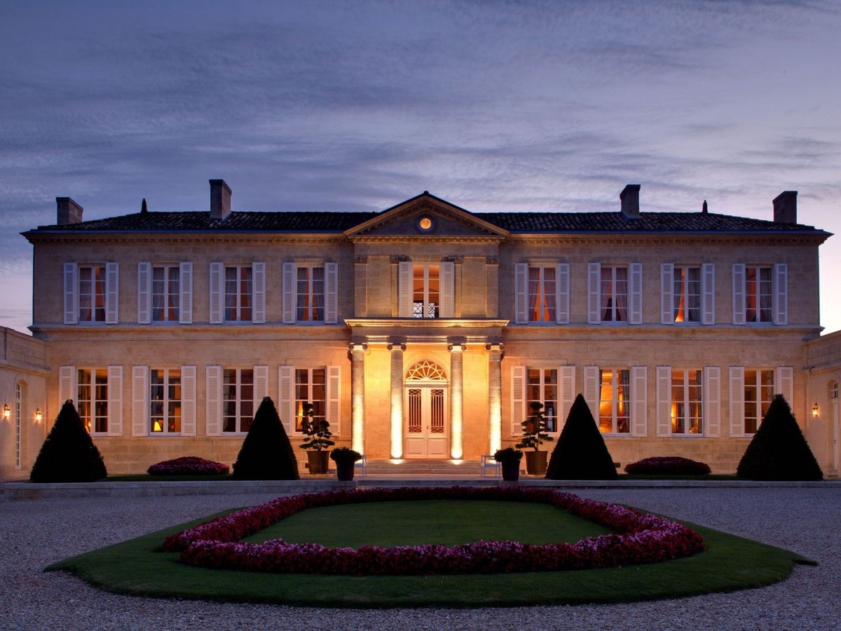 An illuminated French château exterior at dusk with formal gardens in the foreground, featuring a grand entrance and many windows with white shutters.