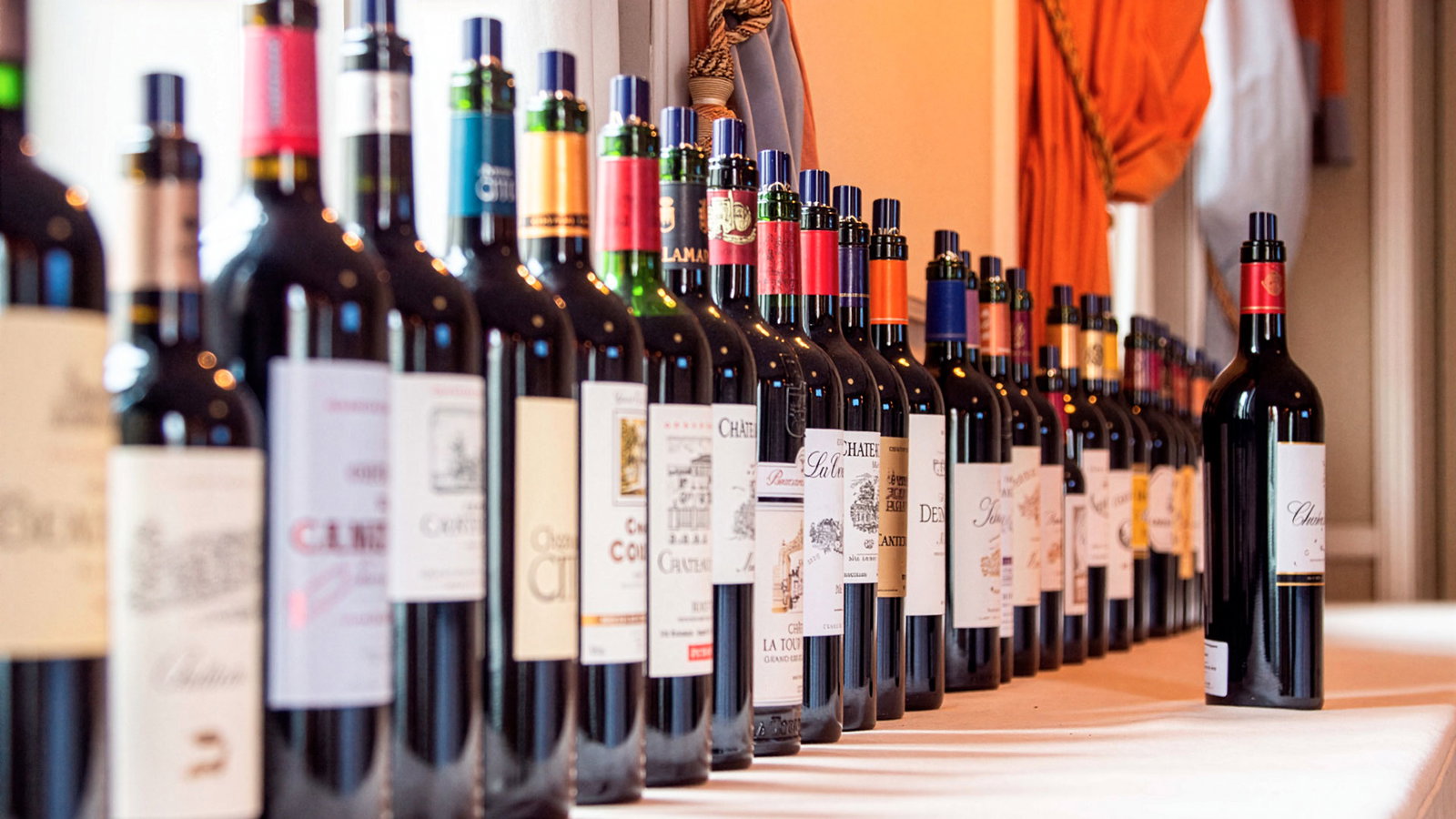 A long diagonal row of 25 to 30 dark Bordeaux wine bottles with varied foil capsules and château labels arranged on a draped tasting table in a warmly lit reception room.
