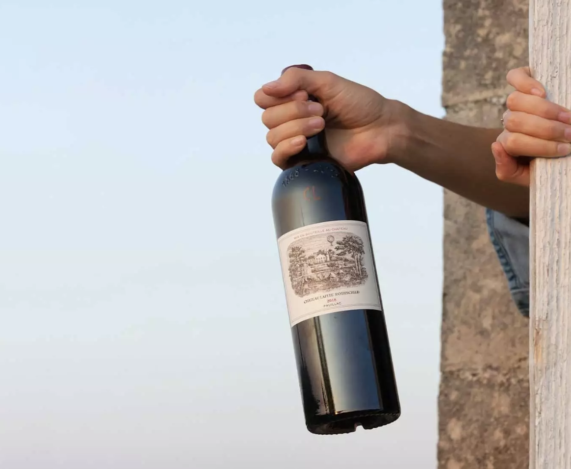 A hand holds a bottle of Château Lafite Rothschild 2018, with the label clearly visible against a light background.