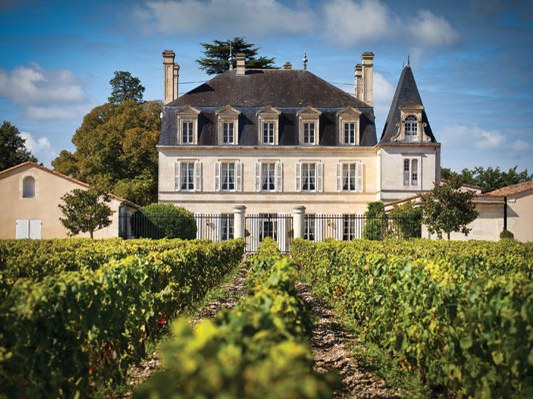 A classic French château, Château Grand-Puy-Lacoste, viewed from between rows of grapevines, showing a two-storey limestone manor with a conical
