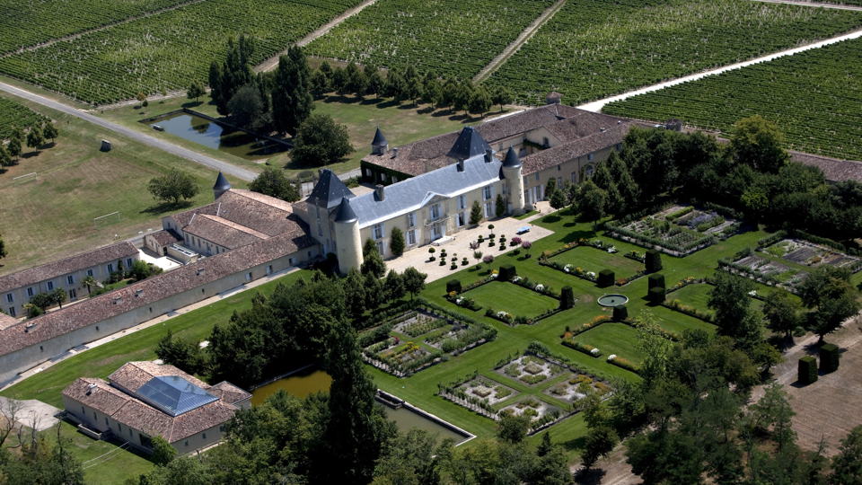 An aerial view of a grand estate with a large, light-colored chateau featuring dark roofs and turrets, surrounded by manicured gardens and extensive