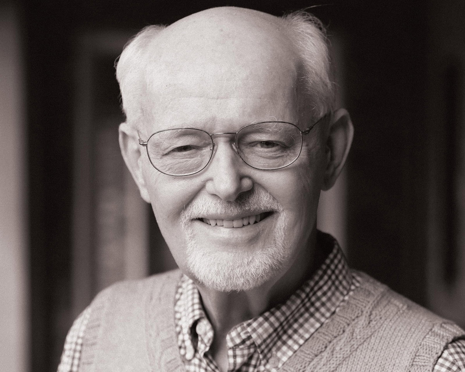 A close-up, black and white portrait shows Mike Veseth, a man with glasses and a white beard, smiling directly at the camera.