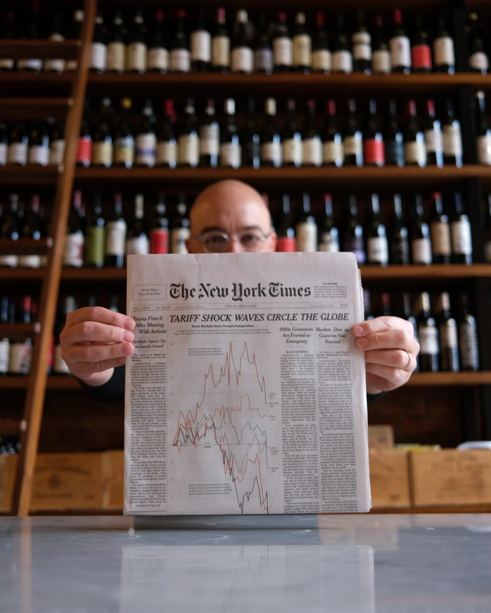 Man holding the April 4, 2025 New York Times with headline "Tariff Shock Waves Circle the Globe" inside a wine shop lined with imported bottles on wooden shelves.