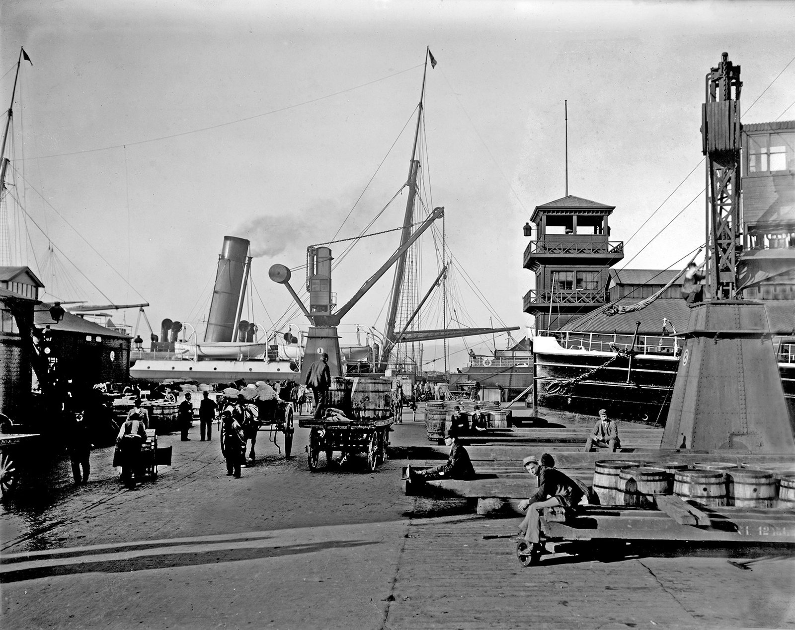 A bustling black and white scene at a port with large ships, cranes, and horse-drawn carts. People are working and resting on the dock.