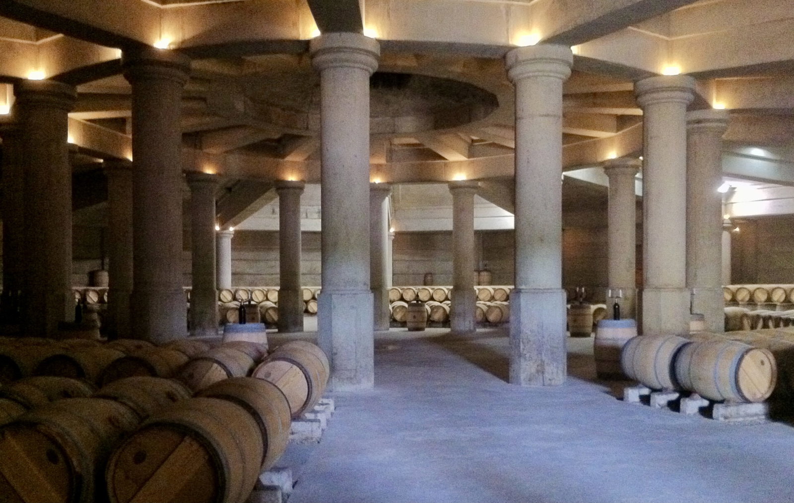 A large, dimly lit wine cellar with rows of wooden barrels stacked on the floor and supported by numerous stone columns.