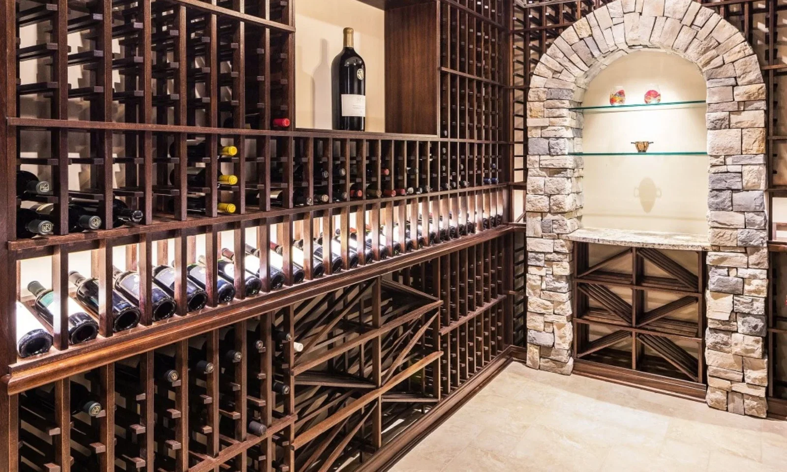 A large, dark wood wine rack filled with numerous bottles of wine, with a stone archway on the right containing shelves and decorative items.