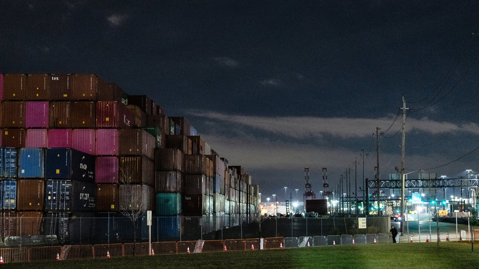 A large stack of shipping containers in various colors, including brown, pink, and blue, under a dark night sky.