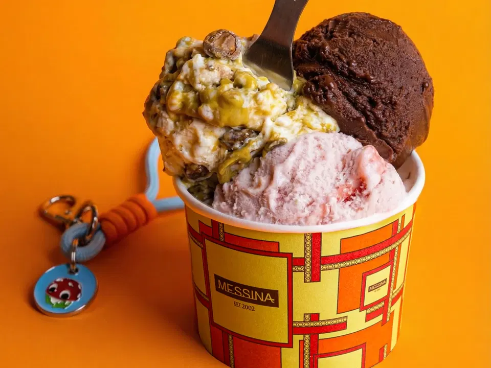 A cup of Messina gelato with three scoops of different flavors: a light green and white swirl with nuts, a dark brown chocolate, and a light pink