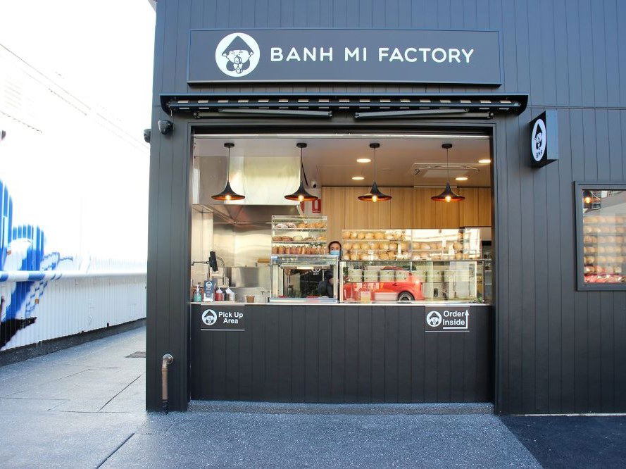 The exterior of Banh Mi Factory, a dark-colored building with a large open window revealing the counter, kitchen, and a person inside.