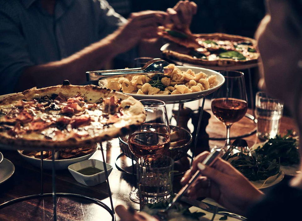 A close-up shot of a table laden with food and drinks, including two pizzas, a plate of fried calamari, and several glasses of wine and water. People