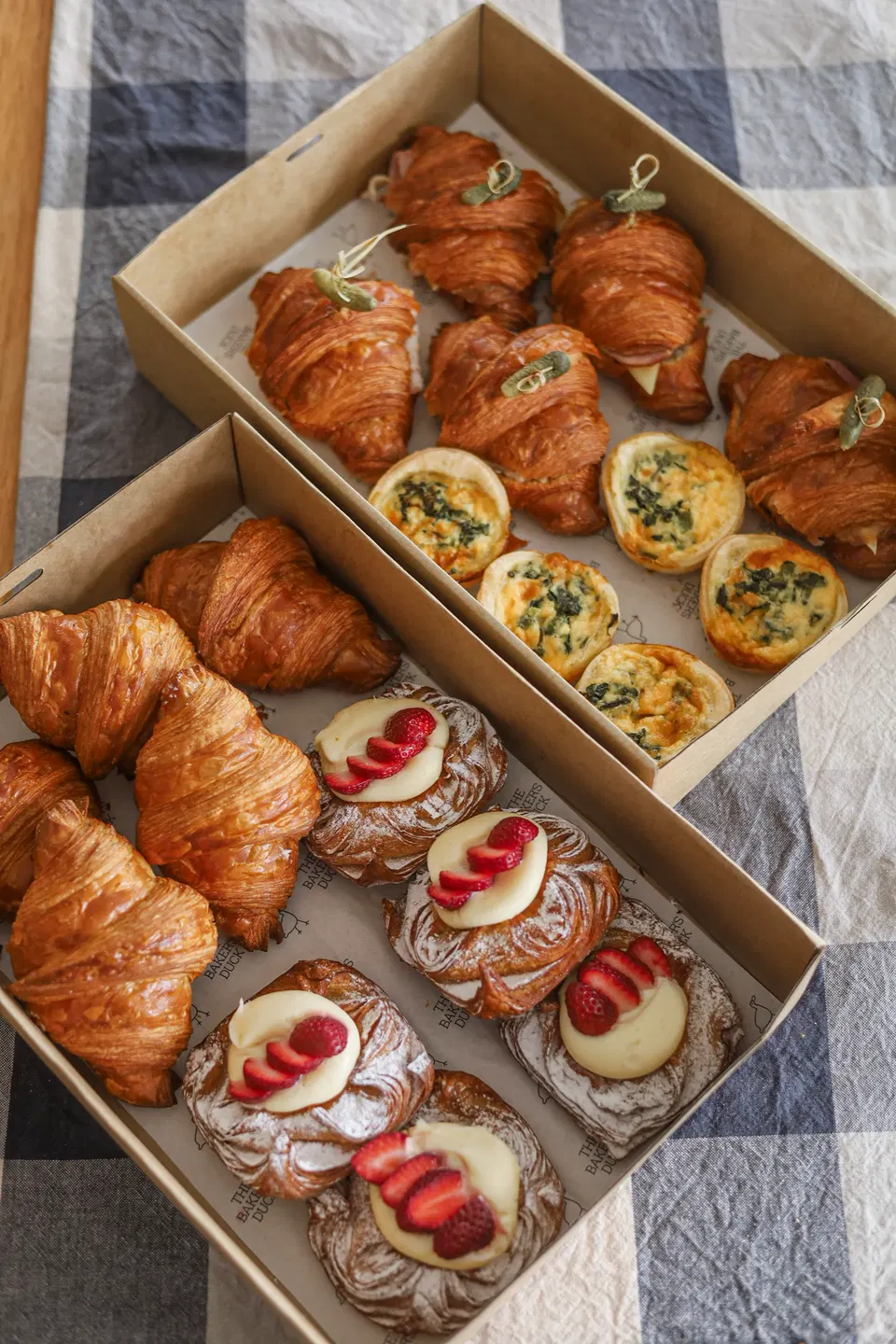 Two cardboard boxes filled with various pastries, including croissants, mini quiches, and danishes topped with cream and strawberries.