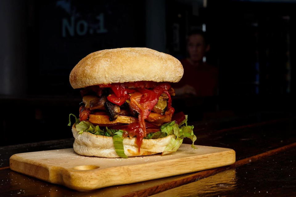 A tall vegetable burger with roasted red peppers, mushrooms, lettuce, and dripping red relish in a round bun, served on a small wooden cutting board.