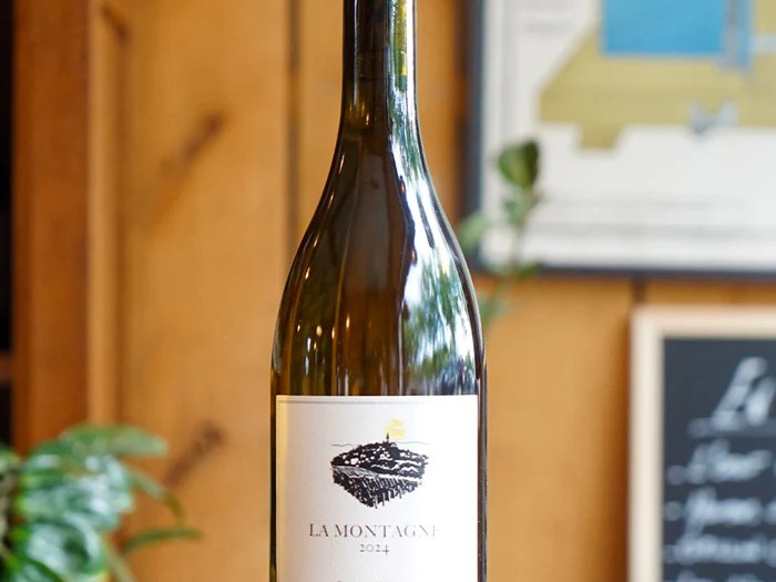 A bottle of La Montagne 2024 by Bastian Wolber, a white wine, stands on a wooden surface in a room with a blurred background.