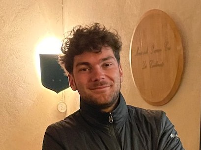 A young man with curly hair, identified as Charles Boigelot, stands with crossed arms in a wine cellar surrounded by oak barrels and wine bottles.