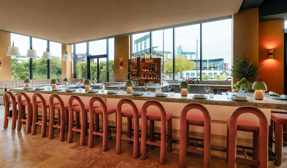 Long marble bar counter with arched terracotta velvet stools inside Via Aurelia restaurant, with Oracle Park stadium visible through floor-to-ceiling windows.