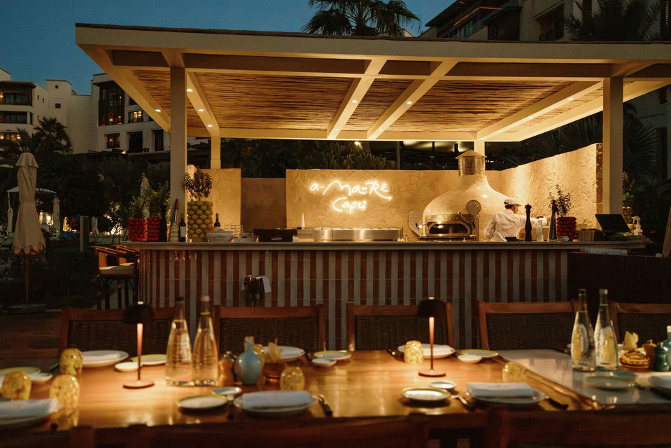 An outdoor restaurant at night with a long wooden table set with plates and glasses, illuminated by small lamps. In the background, a bar area