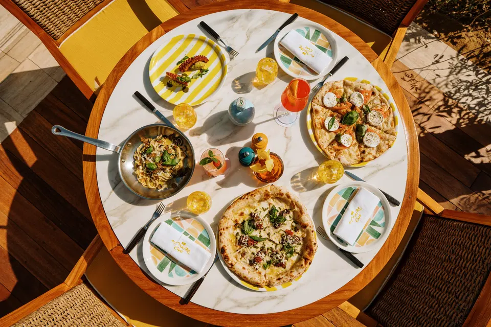 An overhead shot of a marble table set with two pizzas, a pasta dish in a pan, a grilled octopus dish, and various drinks.