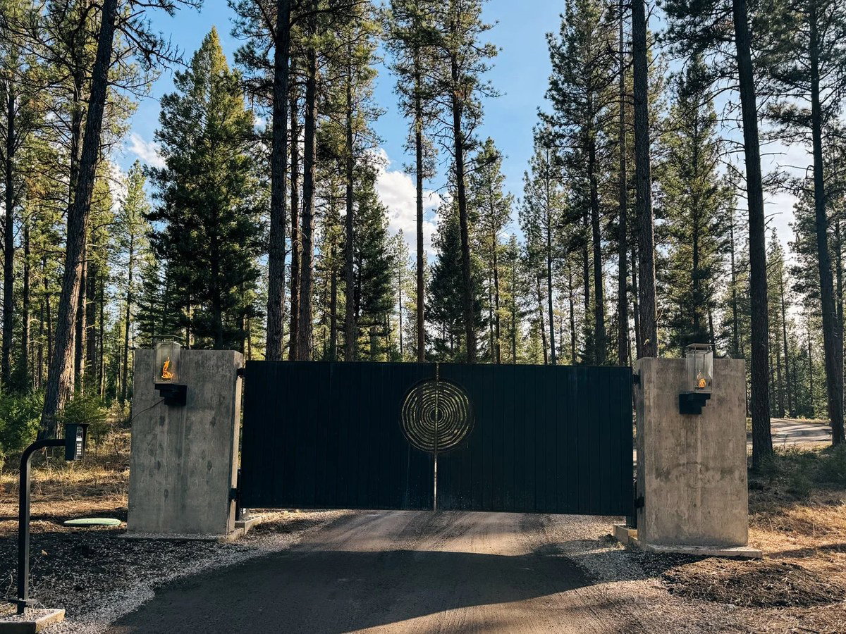 A dramatic black metal gate with a gold concentric-circle medallion, flanked by concrete pillars with flame lanterns, at the end of a gravel driveway