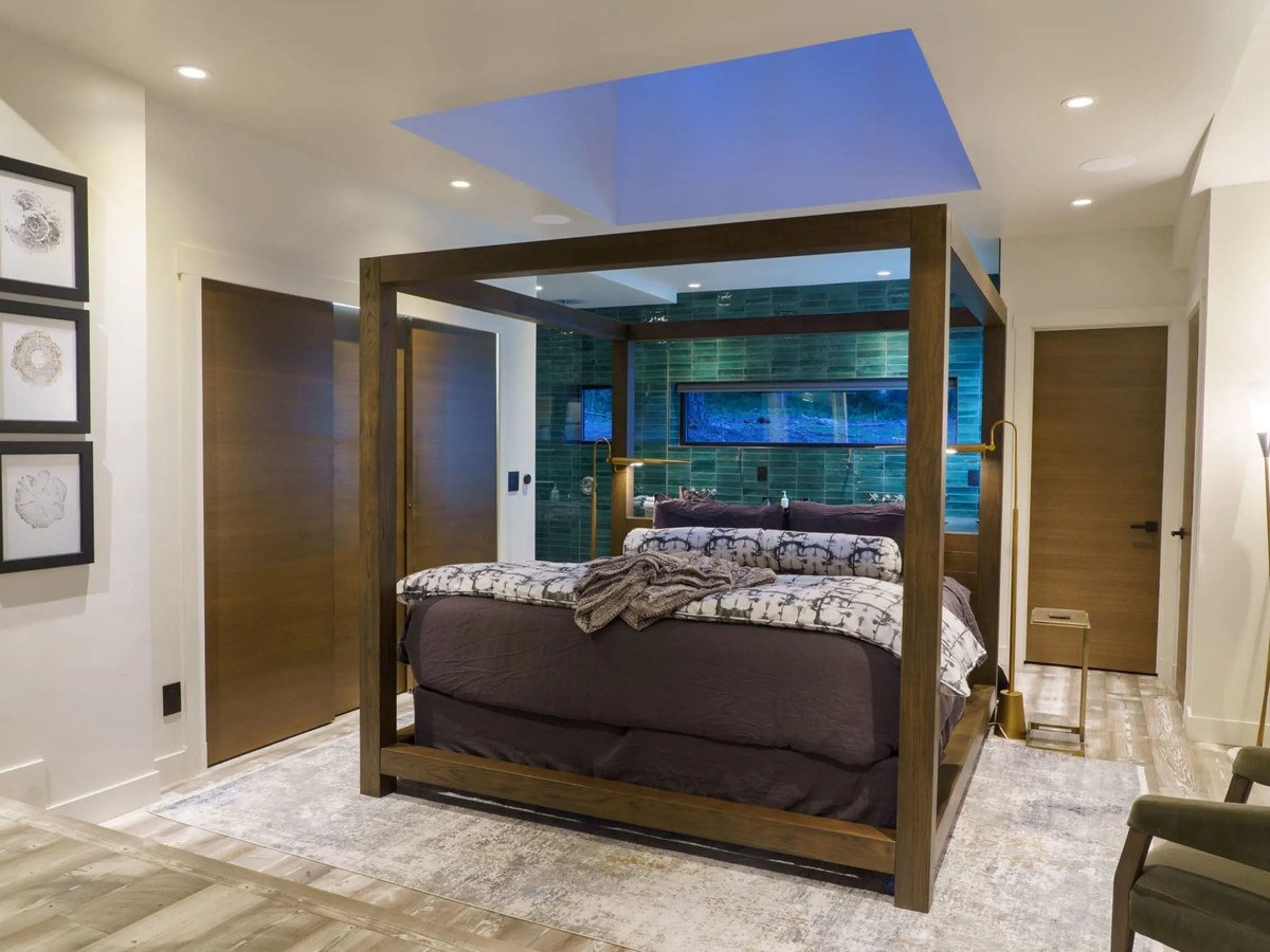 A modern bedroom with a four-poster canopy bed, dark wood frame, and purple bedding, featuring a blue-lit tray ceiling and green tiled wall.