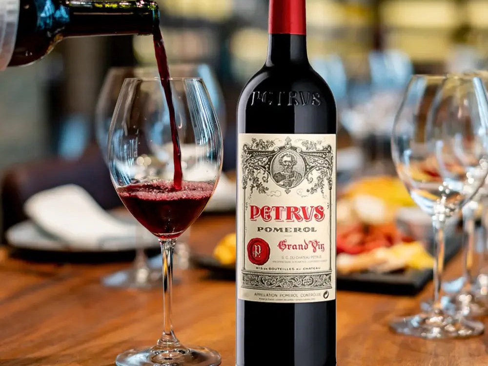 A bottle of Pétrus Pomerol Grand Vin with its distinctive label is shown next to a large Burgundy-style glass being poured with deep garnet red wine.