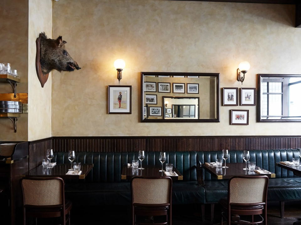 An elegantly appointed restaurant dining room with dark green leather banquette seating, dark wood panelling, and framed prints.