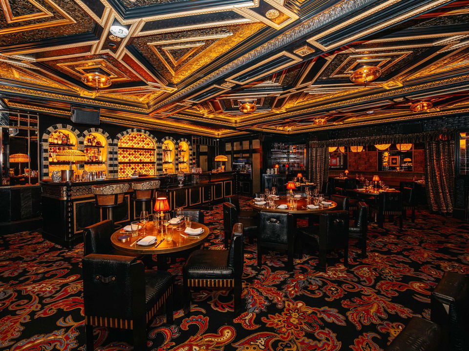 A luxuriously decorated private dining room with an ornate gold and black coffered ceiling, patterned carpet, and an arched bar.