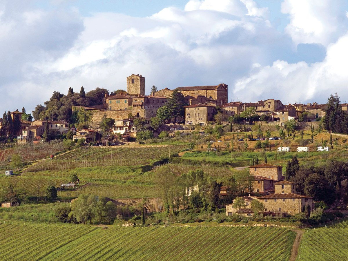 A medieval hilltop village in Tuscany, Italy, surrounded by terraced vineyards under a cloudy sky.