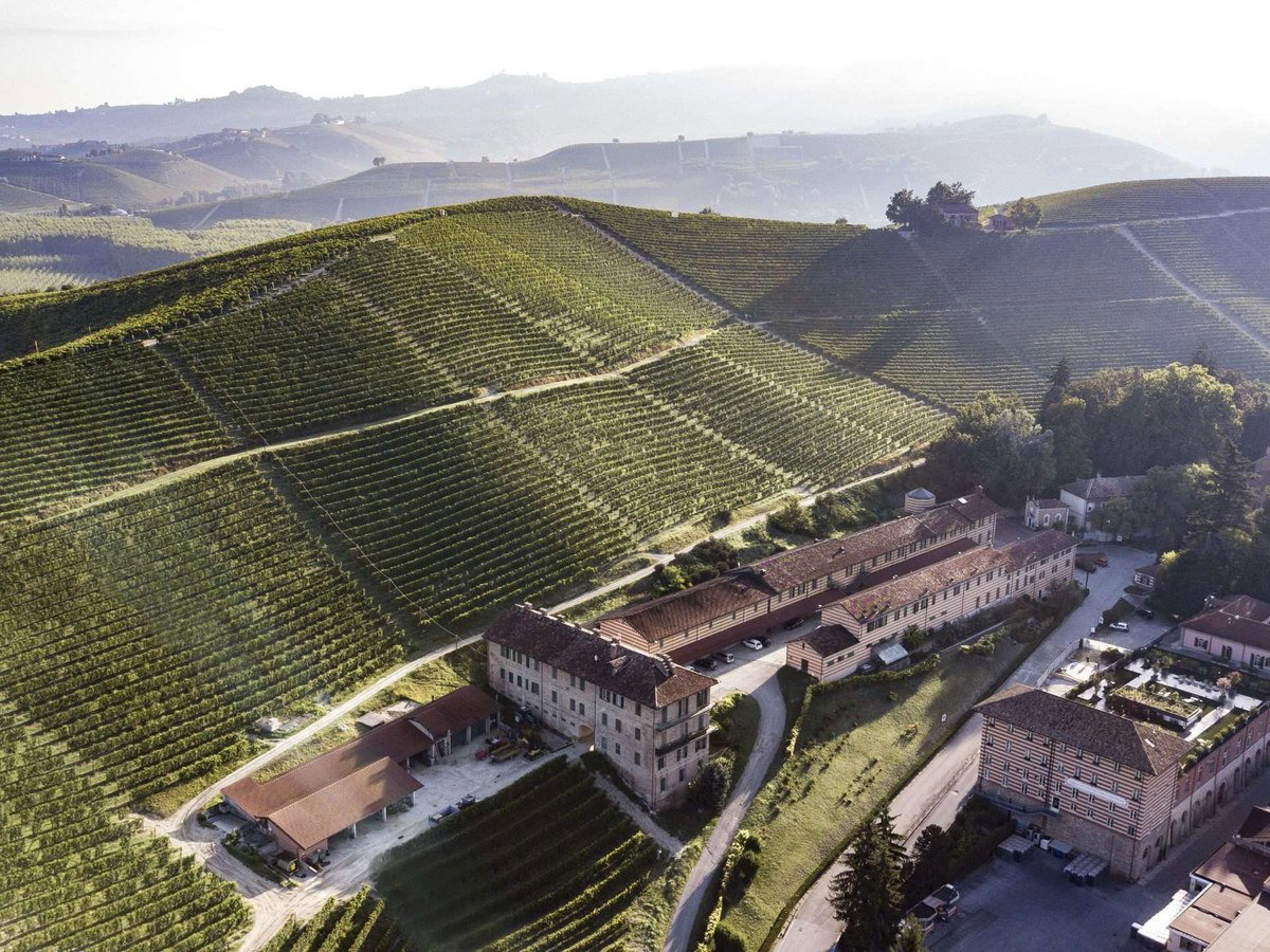 Aerial photograph of a large stone winery building surrounded by manicured vineyard rows on steep hillsides, with misty mountains visible in the distance.