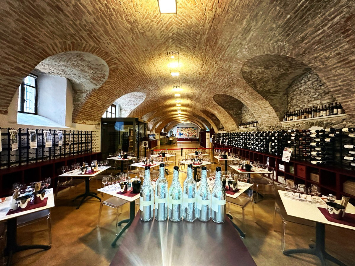 A long, vaulted brick-ceiling room with rows of wine bottles on shelves along the walls. Tables are set with wine glasses, and several foil-wrapped.